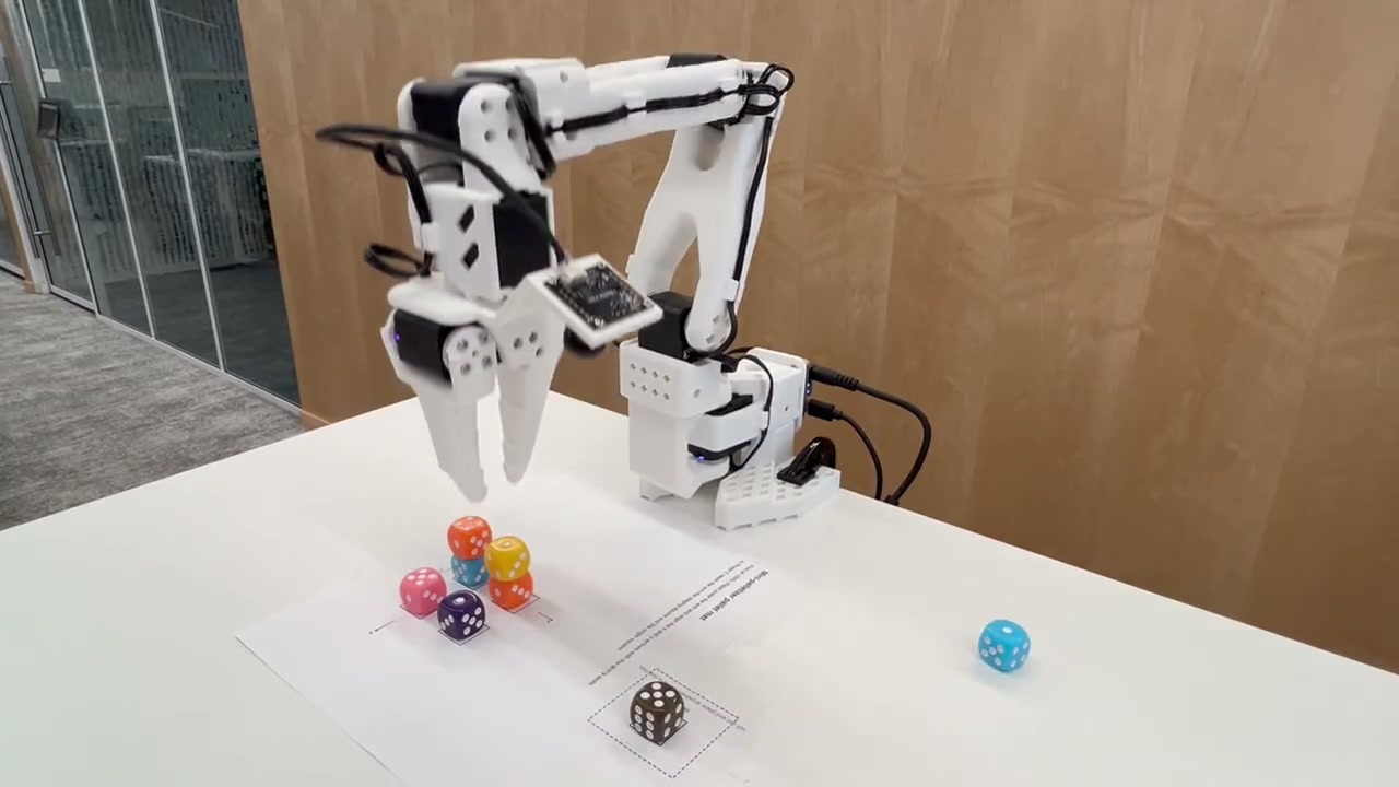 An SO-ARM101 arm on a desk carrying a die toward a partly built stack of dice on a printed pallet mat, with the staging square in the foreground.